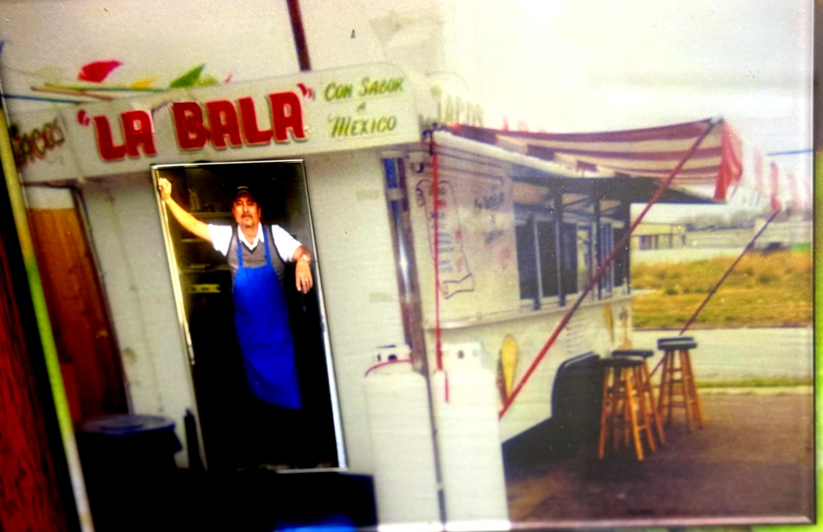 Framed wall photos: the founder in a blue apron in the trailer doorway under the La Bala sign, and the trailer parked in an open lot with a striped awning and wooden stools